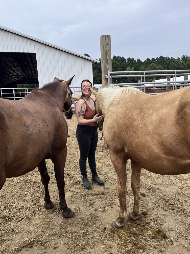 Erin is standing in front of a barn next to two horses and she is smiling very big.