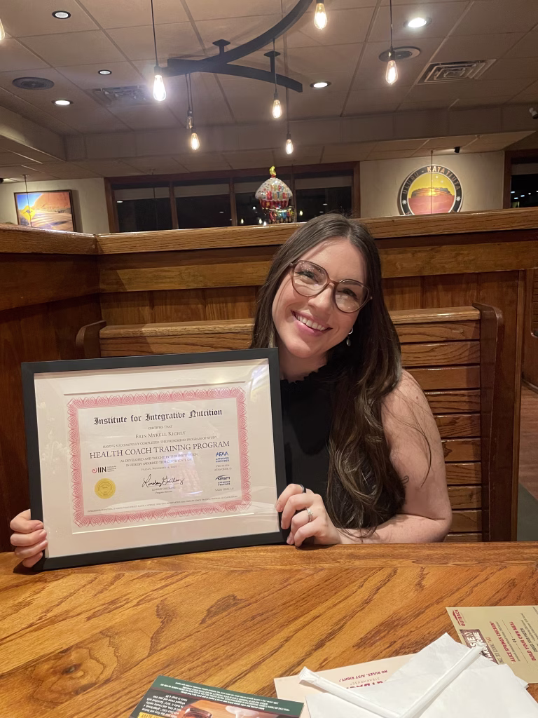 Erin sitting in a dining booth at a restaurant holding her IIN Certificate of completion.